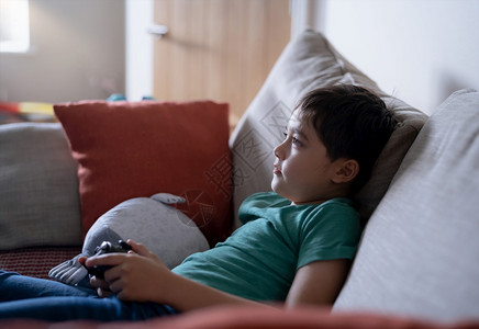 在网上玩电子游戏的男孩Candid拍摄快乐的孩子坐在沙发上拿着游戏控制台坐在沙发上肖莱特孩子在周末家休息时脸朝着看松弛年轻的观图片