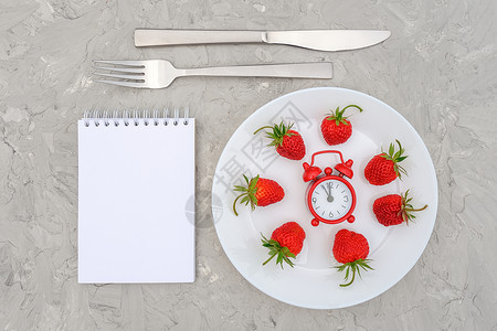 白盘上的红熟草莓浆果餐具红色闹钟和灰石头背景桌上的空白记事本顶视图平躺模型概念饮食和排毒时间或夏季菜单白盘上的红熟草莓浆果色闹钟图片