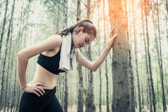 运动型美丽亚洲女在森林里慢跑累了靠在树上毛巾和汗水元素运动和健康概念慢跑和步概念放松和腰痛主题户外活动擦拭图片