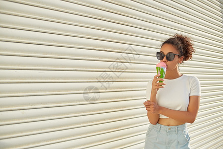 女士幸福夏天穿着白色T恤和轻牛仔裤的年美籍黑人女孩在日光照耀时用冰淇淋抵挡着一堵轻墙吃冰淇淋图片