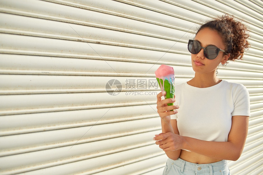 幸福愉快甜点穿着白色T恤和轻牛仔裤的年美籍黑人女孩在日光照耀时用冰淇淋抵挡着一堵轻墙吃冰淇淋图片