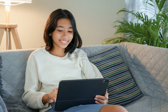 情感触碰美丽的快乐亚洲女孩在晚上坐客厅沙发时使用平板数码工具图片