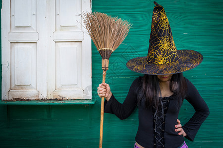 假期农村万圣节艺术设计美丽的年轻女孩戴着巫帽子和扫帚在她手中绿色墙背景木头图片