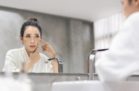 白浴衣的亚洲压力女看着脸朝镜子中的小酒窝试图挤压它担心年轻美丽的女孩在浴室皮肤护理概念中切除她的肛门泡沫康复亚洲人图片