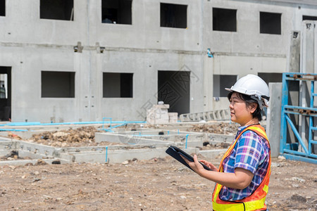 快乐的亚洲女土木建筑工程或设计师身着头盔和安全背心工作并拥有一台触摸不定的平板电脑以查看建筑物或工地的蓝图或计划建筑工地的女程师图片