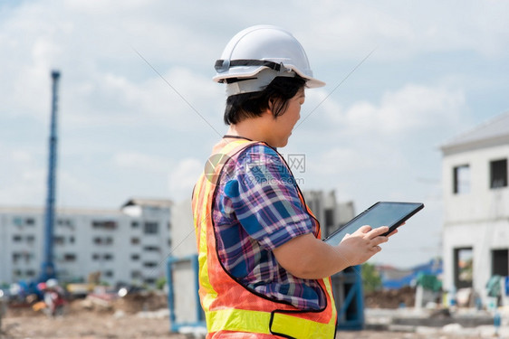 亚洲女土木建筑工程或设计师身着头盔和安全背心工作并拥有一台触摸不定的平板电脑以查看建筑物或工地的蓝图或计划建筑工地的女程师行业在图片