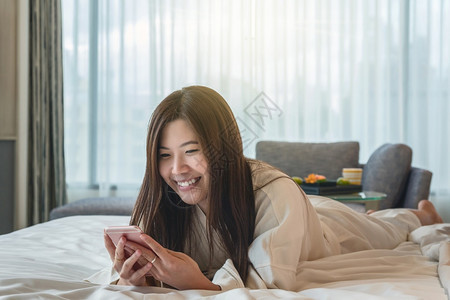 浴绳穿着衣的亚洲妇女使用智能手机在豪华酒店生活方式和休闲概念中醒来时在床上微笑地着行动白色的温泉图片