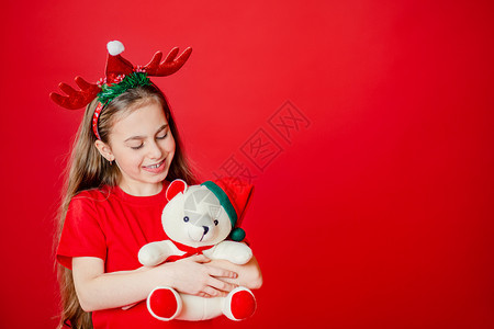 手势包装好的一个有趣快乐女孩肖像头上缠着角绷带抱一只泰迪熊穿着圣诞睡衣背景为鲜红色头抱着一只泰迪熊与鲜红色背景隔开的图片