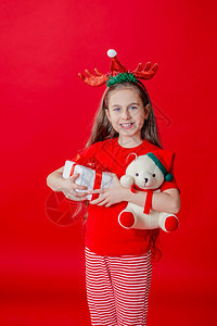 玩具鹿问候一个有趣的快乐女孩肖像头上缠着角绷带抱一只泰迪熊穿着圣诞睡衣背景为鲜红色头抱着一只泰迪熊与鲜红色背景隔开图片