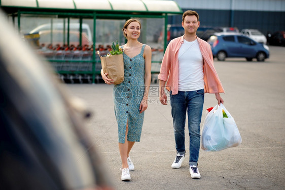 在超市停车场的汽上装有纸袋的年轻家庭夫妇快乐客户从购物中心携带买品车身背景的辆在超市停时装满袋子的年轻夫妇丈中央人们图片