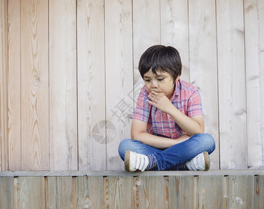坐在木墙边面悲哀的不快乐男孩独自孤单满目沉思地仰望着不安儿童无趣的孤独儿童健康概念的孤独男孩情感肖像危机麻烦强调图片
