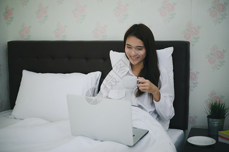 坐着亚洲女人喝咖啡用笔记本在床上工作她笑着享受在家工作的生活互联网笔记本电脑图片