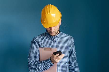 工作业的年轻程师或建筑承包商身穿黄色防护头盔和衬衫站在蓝墙背景前的色面前手持智能电话和剪贴板阅读信息或报告的青年工程师或建筑承包图片