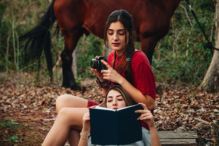 红色的流浪癖微笑年轻一对身着洋装的年轻女子正在阅读一本书森林里用旧相机和马拍照图片