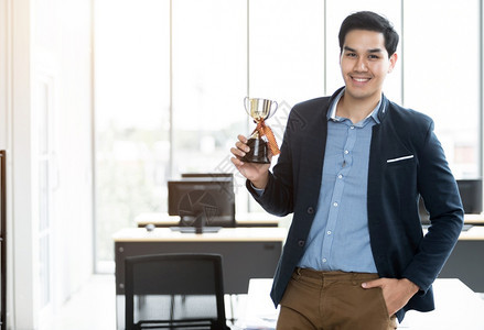 自信的最好幸福成功感觉胜利者肖像尖叫着英俊的年轻亚洲商人在办公室背景中拿着冠军杯在办公室背景图片