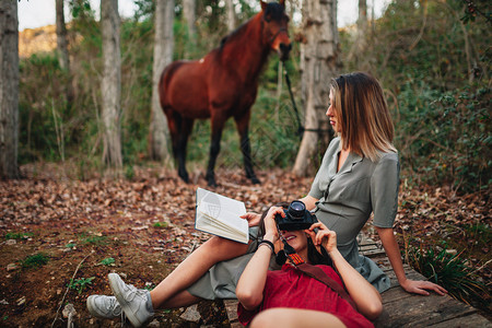 朋友们灰色的年轻一对身着洋装的年轻女子正在阅读一本书森林里用旧相机和马拍照假期图片