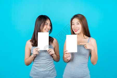 空白的两个笑亚洲青年妇女展示和关于孤立蓝色背景的海报在夏季室内工作穿防弹衣小册子和白广告纸等夏季室内工作登机模型图片