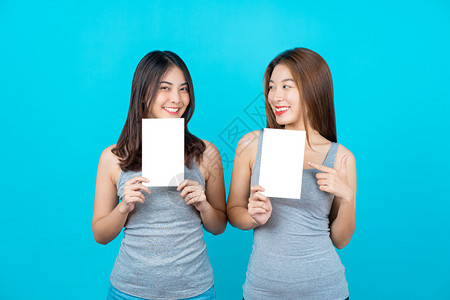 两个笑的亚洲青年妇女展示和关于孤立蓝色背景的海报在夏季室内工作穿防弹衣小册子和白广告纸等夏季室内工作商业摆姿势复制图片