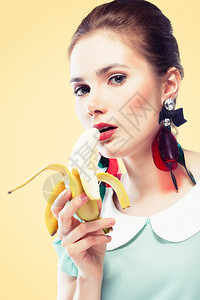 年轻美女嘴唇和眼箭红色的美容粉辣妹化妆时戴漂亮的塑料耳环吃香蕉黄色背景针纹风格拉普捷夫感的白种人图片