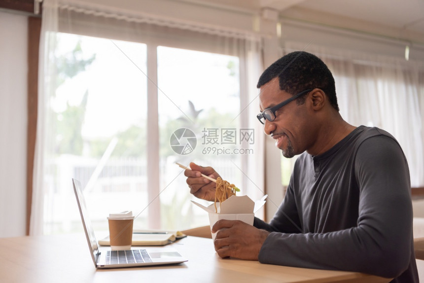 桌子非裔美国人男在用笔记本电脑打视频话时一边吃即杯面一边吃筷子碗面一边笑着黑男手拿沙巴从盒子里取走的眼镜一边在家里吞食19号隔离图片