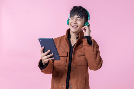 亚洲快乐一位在耳机上英俊的年轻男子在听音乐用平板电脑对侧眼双看镜头着爱中隔绝的相机拍着粉红色空白间工作室背景成人快乐的游戏图片