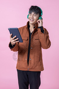 吸引人的一个亚洲男子英俊的年轻耳机正在监听音乐用平板电脑对着侧眼的睛看着爱中孤立的相机在粉红空白复制间室背景上年轻的潮人图片