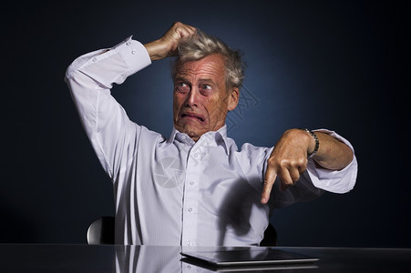 不开心情绪非常激动沮丧的老年商人用平板电脑指着桌子上躺的平板电脑用手撕他的头发注意力撕裂背景图片