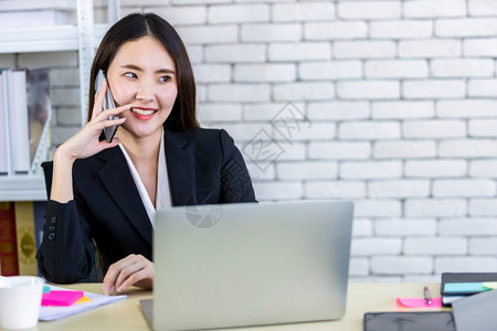 称呼年轻的咖啡成功快乐亚裔年轻女商人抓住手持智能机用笔记本电脑在白色木桌墙办公室背景的白木桌子上工作背景图片
