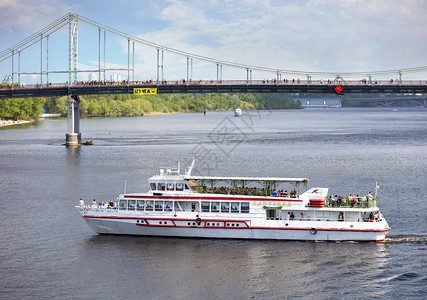 堤在白色游乐船上人们在城市公园和基辅河对面行人桥的背景下在Dnieper河上度过一个明暑夏日白帆船在阳光明媚的天气中载人穿越Dn图片