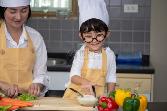 亚洲年轻母亲有儿子和的亚洲女年轻母亲烹饪沙拉食物配菜端着番茄和胡萝卜盘子里放铃椒供幸福家庭煮饭时用在家做享受生活方式午餐勺子为了图片