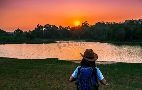 空气成人带着帽子和背包的女旅游者站着心情放松看着池塘和常青林后面的日落独自旅行的年轻女享受美丽的景色夕阳女士图片
