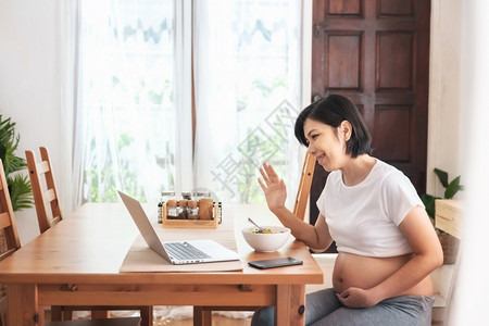 愉快沟通乐笑的亚裔年轻母亲在网上视频电话在她家怀孕时用笔记本电脑聊天和与家人握手时欢乐而美丽的孕妇享受肚子背景图片