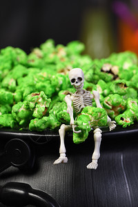 糖果一具尸骨坐在大锅的边缘手里握着一只绿色甜爆米花在万圣节特餐大杂烩死的图片