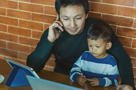 有儿子的亚洲单身父亲正在透过科技笔记本电脑寻找这幅漫画再加上在自学或家庭校和单亲长概念的阁楼里住时电话呼唤着工作技术单身的棕色图片