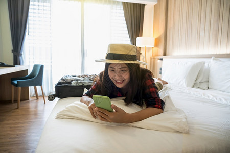 幸福的亚洲旅行者妇女在度假豪华室内卧设计旅游和行概念上睡觉和使用智能手机睡在旅馆或店宿舍卧室的床上并使用智能手机在假日旅行游和概图片