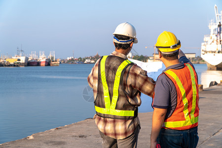 讨论同事两名工程师站在码头着身安全头盔持有蓝图并参考施工构想中的计划即高效施工管理程概念AC54970技术图片