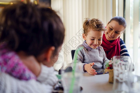聪明的老小男孩caucasiansian小男孩儿子坐在母亲或姑的膝盖上而女朋友正看着她们笑地拿智能手机玩游戏打视频电话家庭游戏年图片