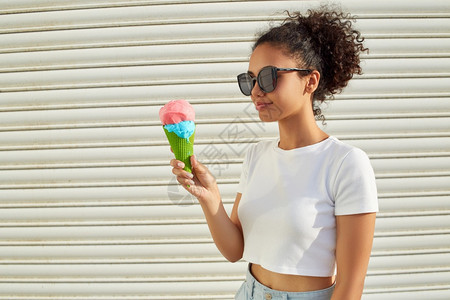穿着白色T恤和轻牛仔裤的年美籍黑人女孩在日光照耀时用冰淇淋抵挡着一堵轻墙吃冰淇淋可口非洲裔热的图片
