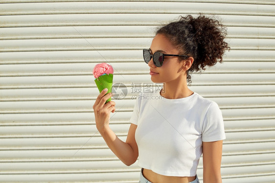 肖像穿着白色T恤和轻牛仔裤的年美籍黑人女孩在日光照耀时用冰淇淋抵挡着一堵轻墙吃冰淇淋甜的年轻图片