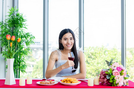 情人节的概念笑亚裔年轻女坐在餐桌食品展上带着酒杯在餐厅背景中盛着的美酒杯裙子红葡萄图片