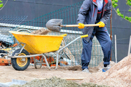 劳动者行业放在活跃的建筑工地背景下一名身穿蓝色工作服和黄手套的工人将沙子装入黄色建筑独轮车复制空间建筑工地的一名人将沙子铲入黄色图片