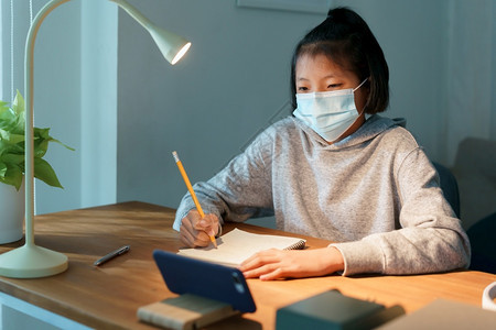 年轻的桌子亚洲女孩正在做家庭作业夜间用智能电话进行网上学习亚洲妇女晚上戴着保护面具的流感行病或在家里使用COVID19健康和疾病图片