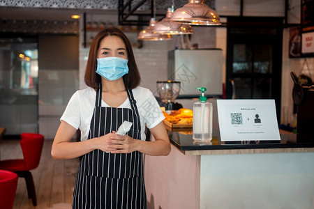 爆发预防身戴面罩和持有红外前额温度计的亚洲女服务员在进入餐馆咖啡店之前检查顾客身上的症状以检查体温在进入餐厅咖啡店之前检查客户的图片