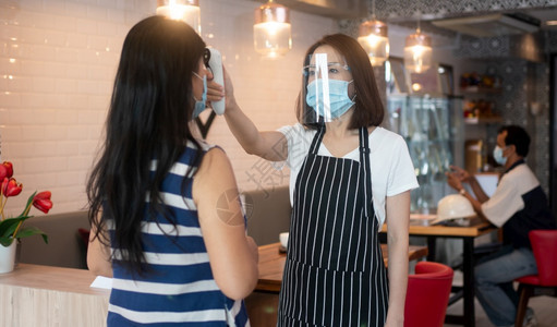 身戴面罩和持有红外前额温度计的亚洲女服务员在进入餐馆咖啡店之前检查顾客身上的症状以检查体温在进入餐厅咖啡店之前检查客户的体温图片