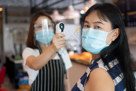 预防电晕身戴面罩和持有红外前额温度计的亚洲女服务员在进入餐馆咖啡店之前检查顾客身上的症状以检查体温在进入餐厅咖啡店之前检查客户的图片
