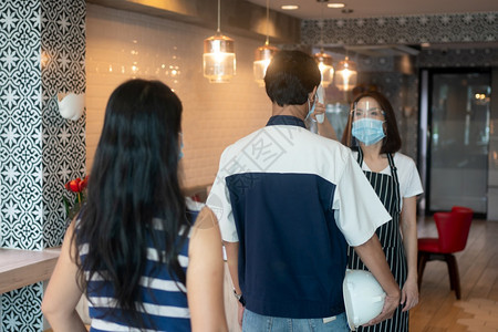 服务器当地的身戴面罩和持有红外前额温度计的亚洲女服务员在进入餐馆咖啡店之前检查顾客身上的症状以检查体温在进入餐厅咖啡店之前检图片