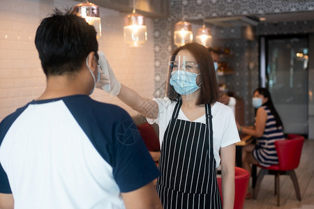 电晕卫生身戴面罩和持有红外前额温度计的亚洲女服务员在进入餐馆咖啡店之前检查顾客身上的症状以检查体温在进入餐厅咖啡店之前检查客户的图片