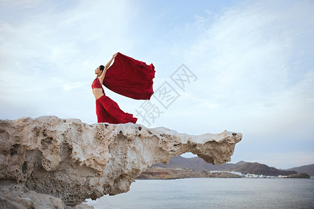 享受摆姿势身穿红礼服的黑发年轻女人在海滨附近岩石上跳舞喜悦图片