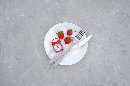 甜的食品白盘上红熟草莓浆果灰色石头背景桌上的餐具和红色闹钟顶视图平躺复制空间模板概念饮食和排毒时间或夏季菜单白盘上的红熟草莓浆果图片