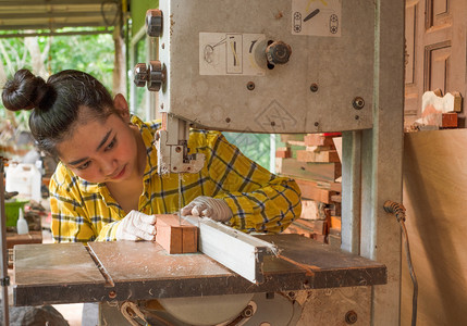 站立妇女正在工作板上用木车间匠机的带锯子发电工具在作台上手艺切割木材人锯板从工人那里经营一家庭企业建造乐器木制的图片